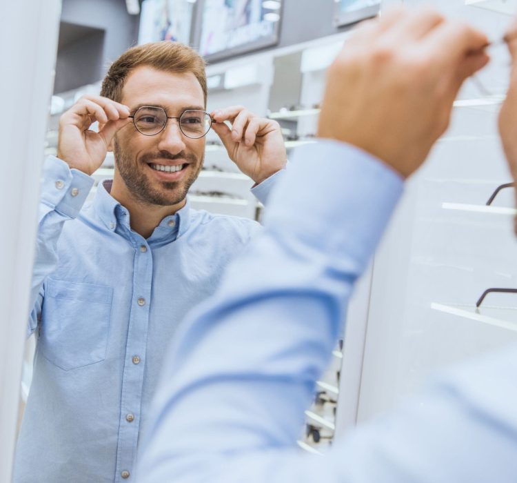 partial view of cheerful young man choosing eyeglasses and looking at mirror in optica